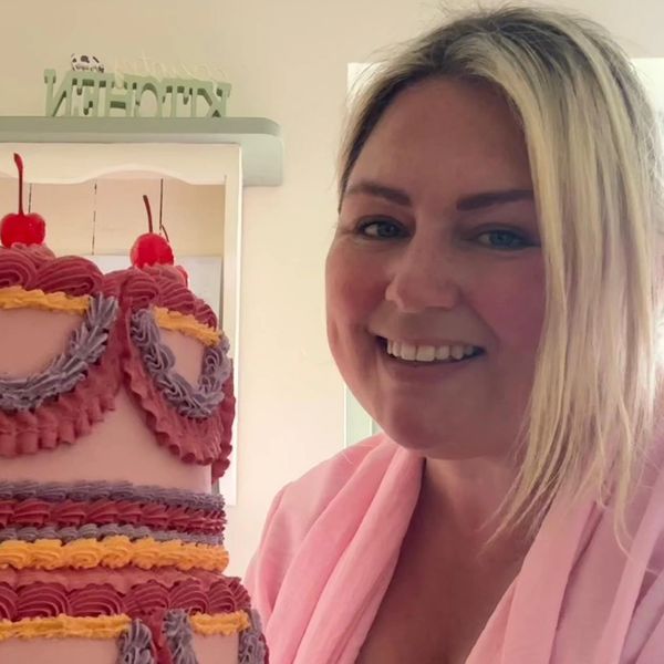 Woman proudly holds a large, intricately decorated three-tier cake with cherries on top.