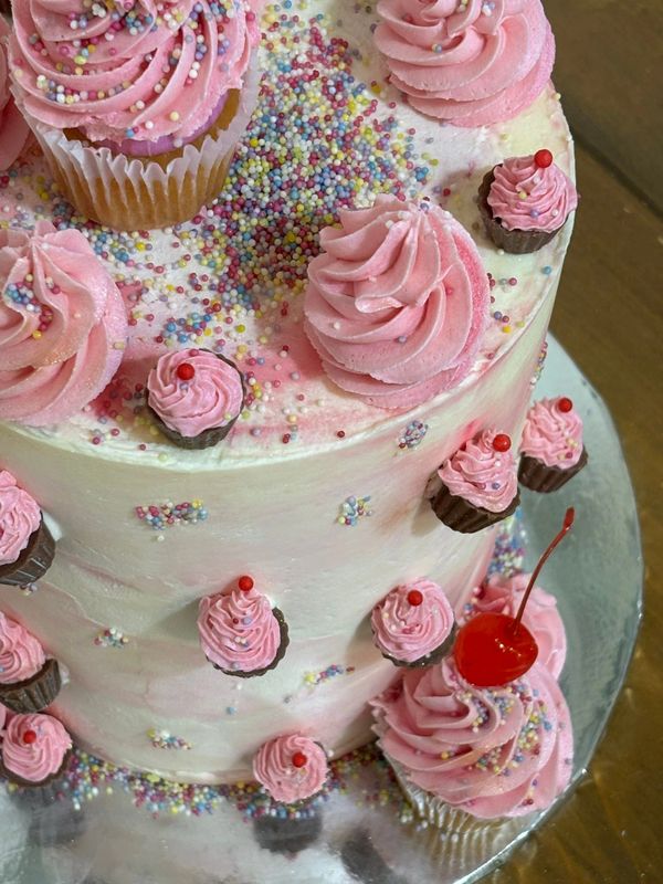 A pink frosted cake decorated with cupcakes, mini cupcakes, sprinkles, and a cherry.