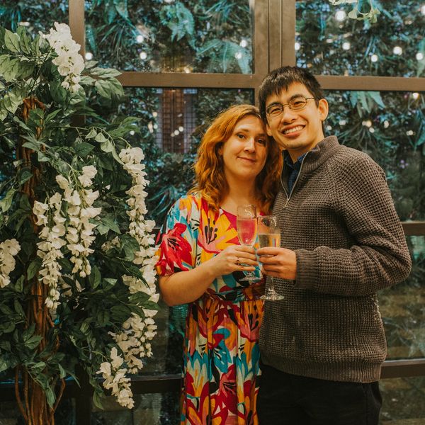 Kate and Alfred posing with a glasses of champagne in front of a floral display.