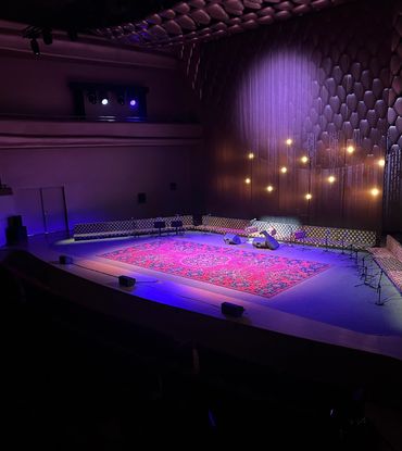 A dimly lit theater stage with a red carpet and a person lying down under a spotlight.