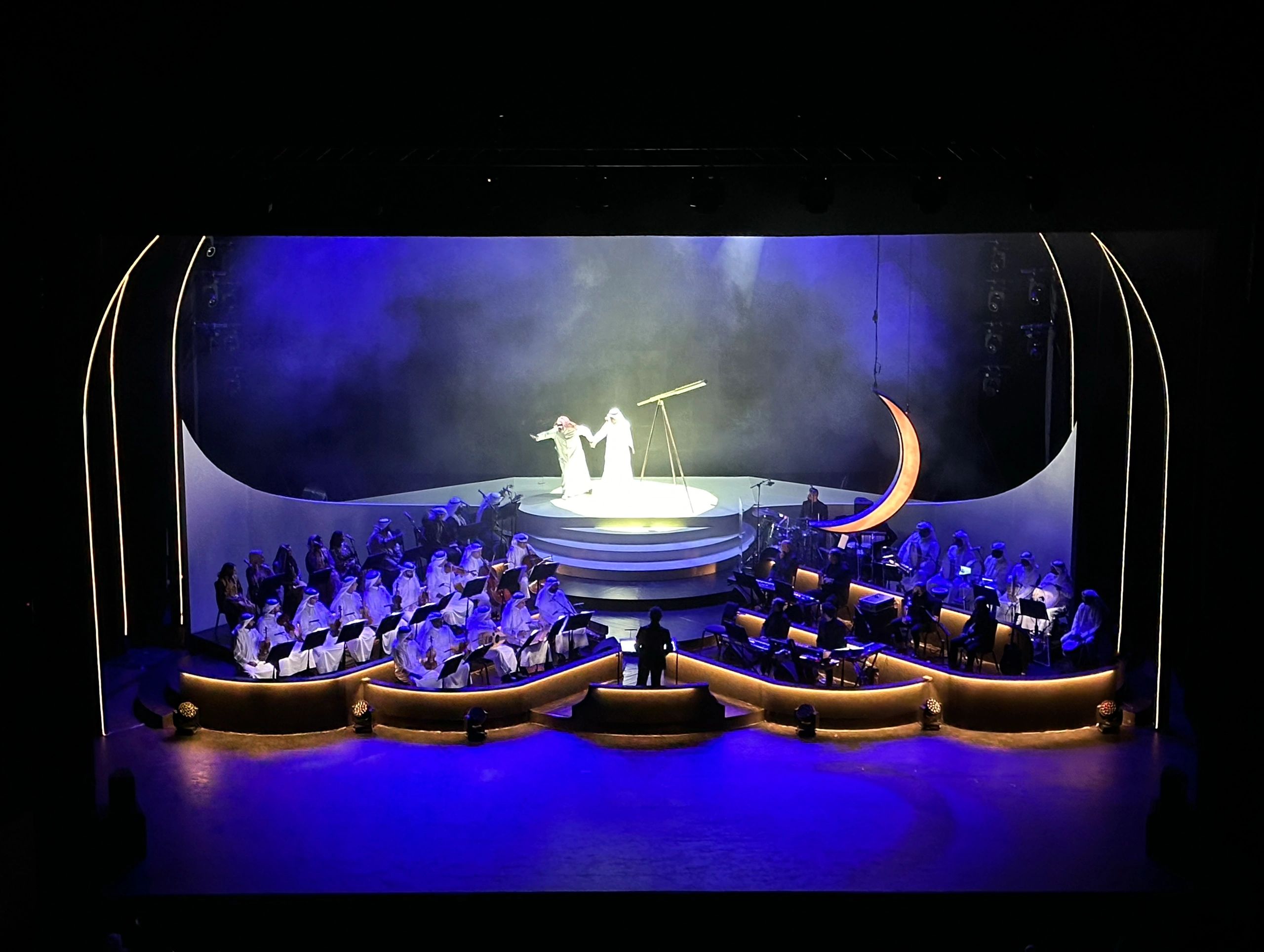 A theatrical performance with musicians, featuring two actors, a telescope, and a crescent moon prop on stage.