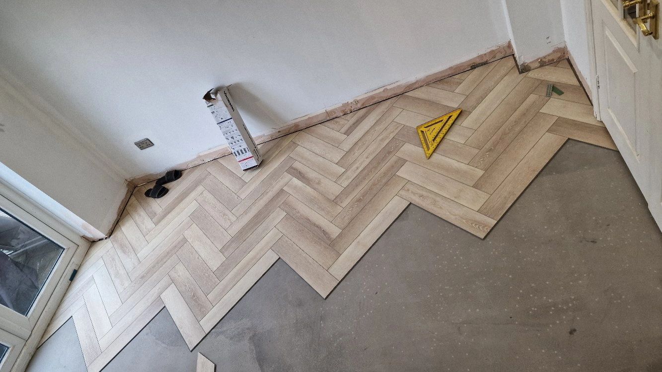 Partially installed herringbone pattern wooden floor with tools and slippers nearby.