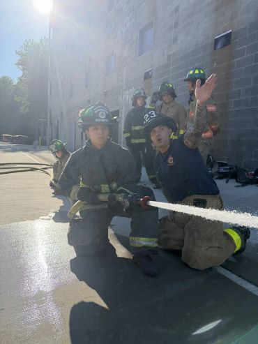 Firefighters training with a hose under sunlight near a building.