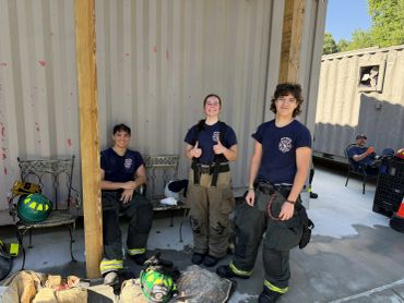 Three firefighters in uniform resting outside a building under a wooden shelter.