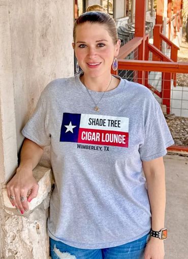 Woman in a gray shirt with 'Shade Tree Cigar Lounge' text, standing by a stone wall.