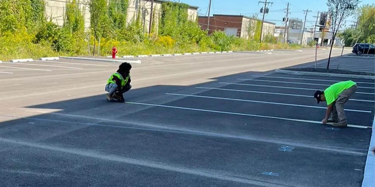 Two workers in neon vests marking lines in an empty parking lot under sunlight.