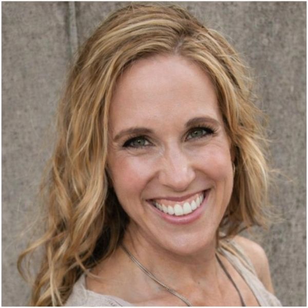 Smiling woman with wavy blonde hair and green eyes posing in front of a textured wall.