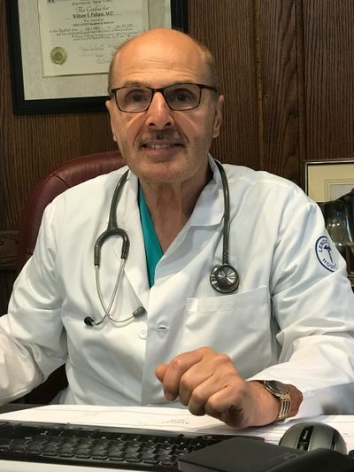 Elderly male doctor in white coat with stethoscope sitting at desk.