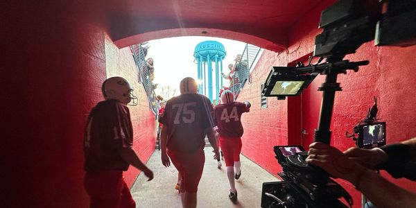 Football players walk through a tunnel toward the field, filmed by a camera crew.