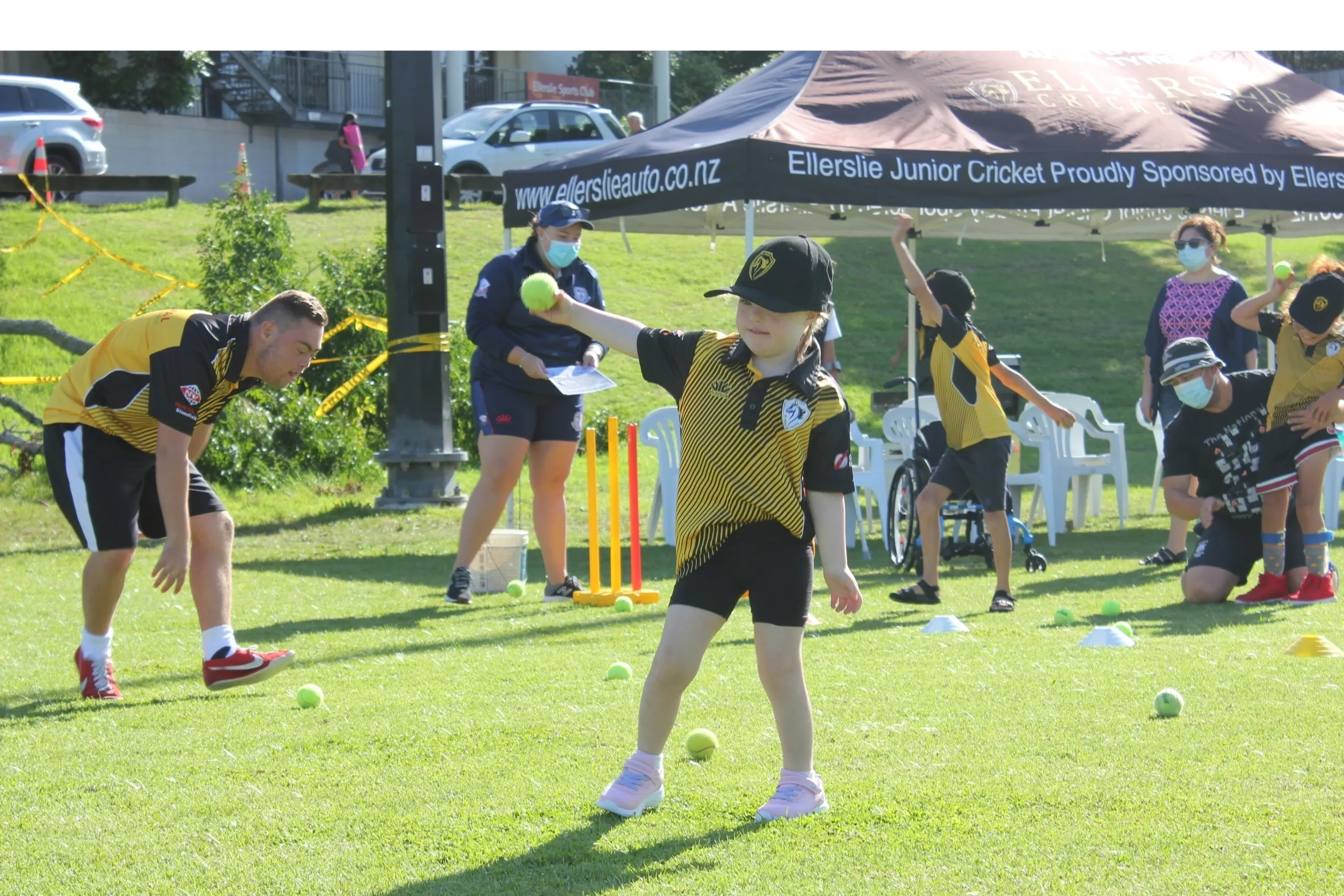 Children in cricket uniforms practicing with coaches on a sunny field.