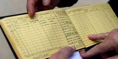 Hands holding and pointing at an open yellow ledger with handwritten entries.