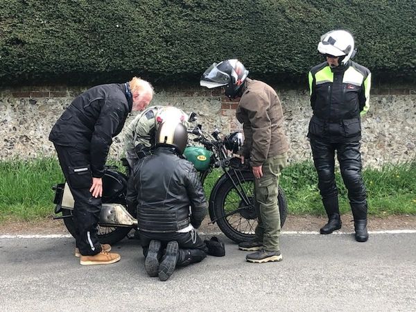 Four motorcyclists inspect a vintage motorcycle on the roadside.