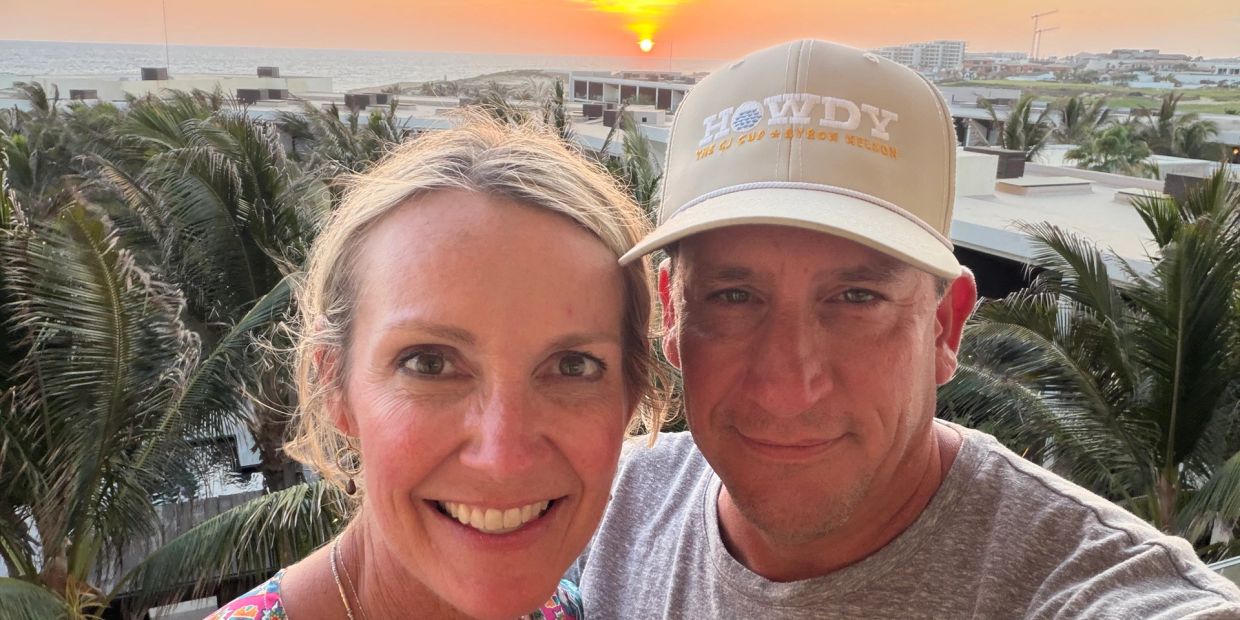 Smiling couple takes selfie at sunset with palm trees and ocean in background.