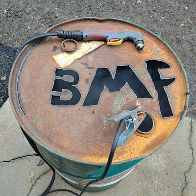 Rusty barrel with 'BMF' cut out and welding tools on top.