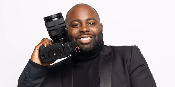 Smiling man in a black suit holding a camera over his shoulder.