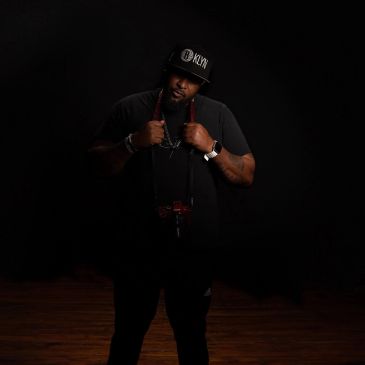 Man posing confidently in dark clothing and a black cap against a dark background.