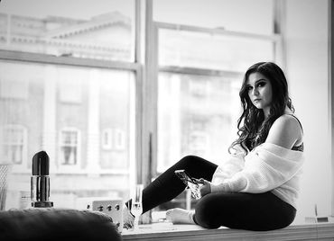A woman sits by a large window holding a glass, deep in thought.