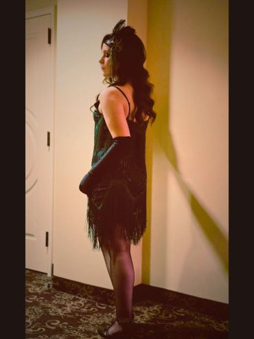 Woman in vintage flapper dress and gloves stands in profile indoors.