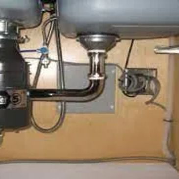 Under-sink plumbing with pipes, disposal unit, and water lines. Some stains on the wall.