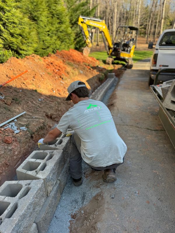 Worker building a stone retaining wall outdoors with machinery nearby.