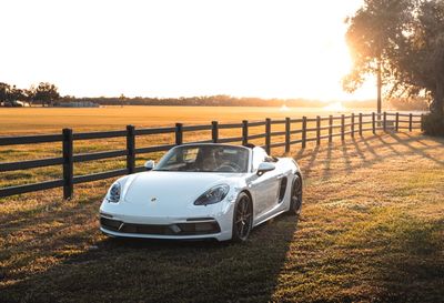White convertible sports car parked on grass at sunset near a wooden fence.