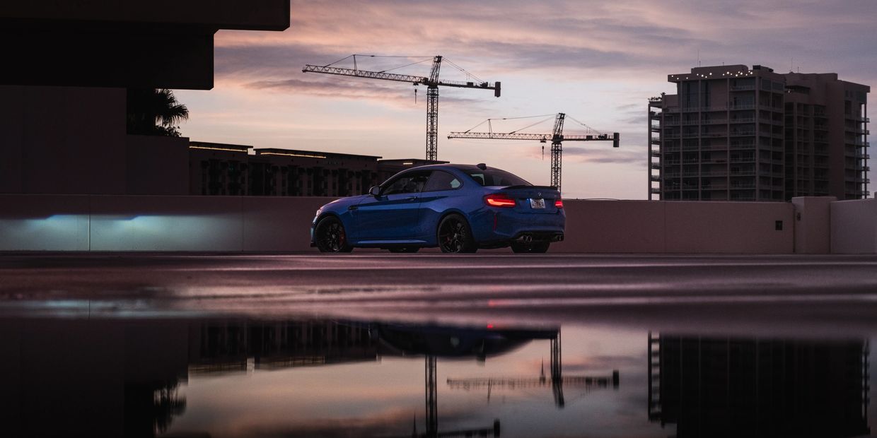 Blue BMW M3 sports car parked with construction cranes and a building at sunset.