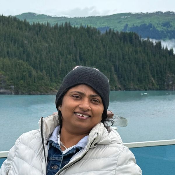 Woman in a winter jacket and beanie smiling by a lake with forested hills.