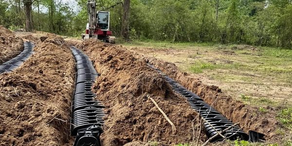 Trenches with drainage pipes being installed in a wooded area.