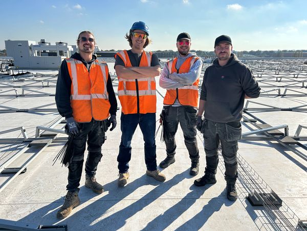 Four construction workers posing on a rooftop with safety gear and orange vests.