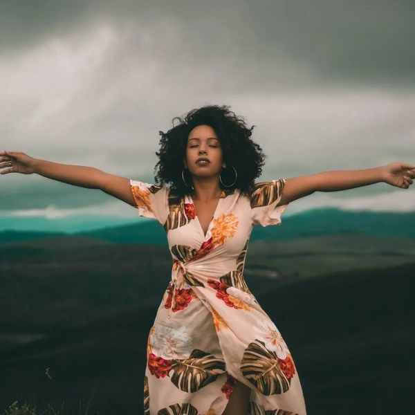 Woman in floral dress with arms outstretched, eyes closed, in a natural outdoor setting.