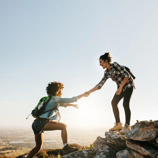 2 women climbing - 1 reaching out to the other to help. Both wearing checked shirts and rucksacks