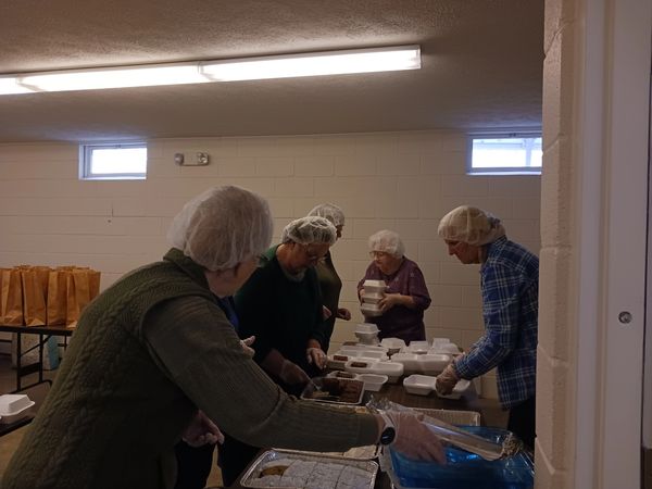 Volunteers serving food in a community kitchen, wearing hairnets and gloves.