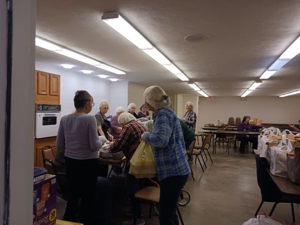 Group of elderly people gathered in a community room with bags and tables.