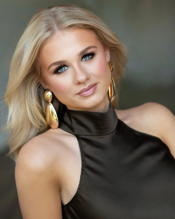 Blonde woman with blue eyes and gold earrings in black dress.