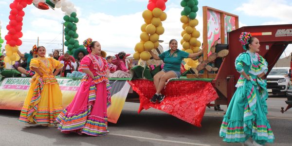 Women in traditional dresses dance at a parade float.