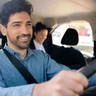 Man smiling while driving a car with a passenger in the back seat.