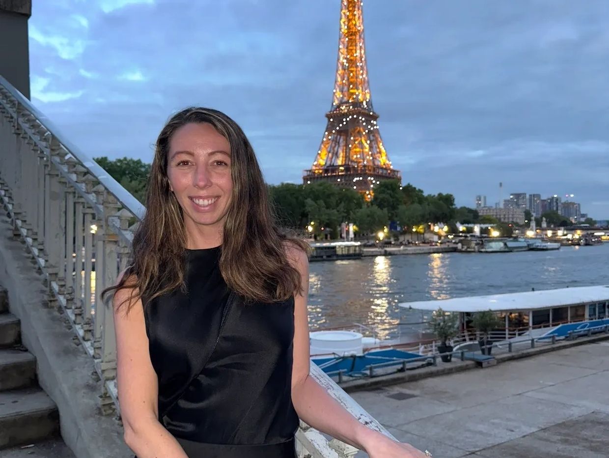 Woman in black dress posing near the Seine with illuminated Eiffel Tower at dusk.