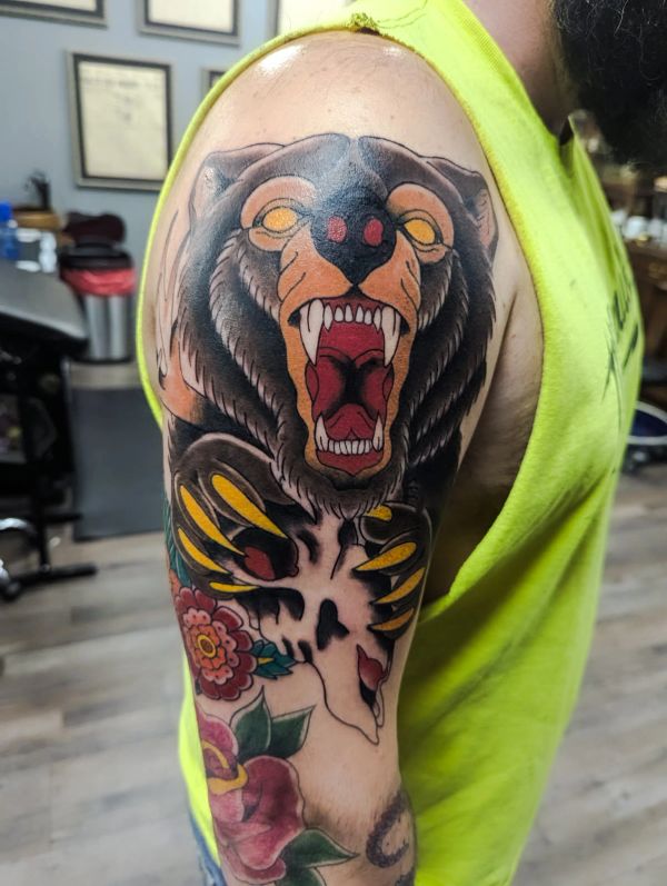 Colorful tattoo of a roaring bear with a skull and flowers on upper arm.