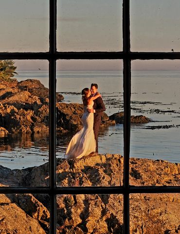 A couple embraces on rocky shore at sunset, viewed through a window.