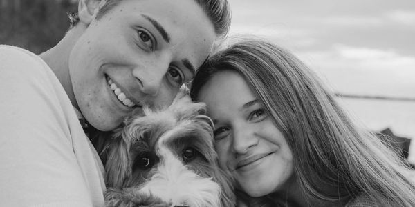 Two people smiling with a dog in a close-up black and white photo.