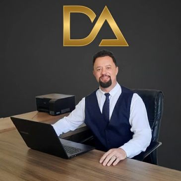 A man in business attire sits at a desk with a laptop, smiling.
