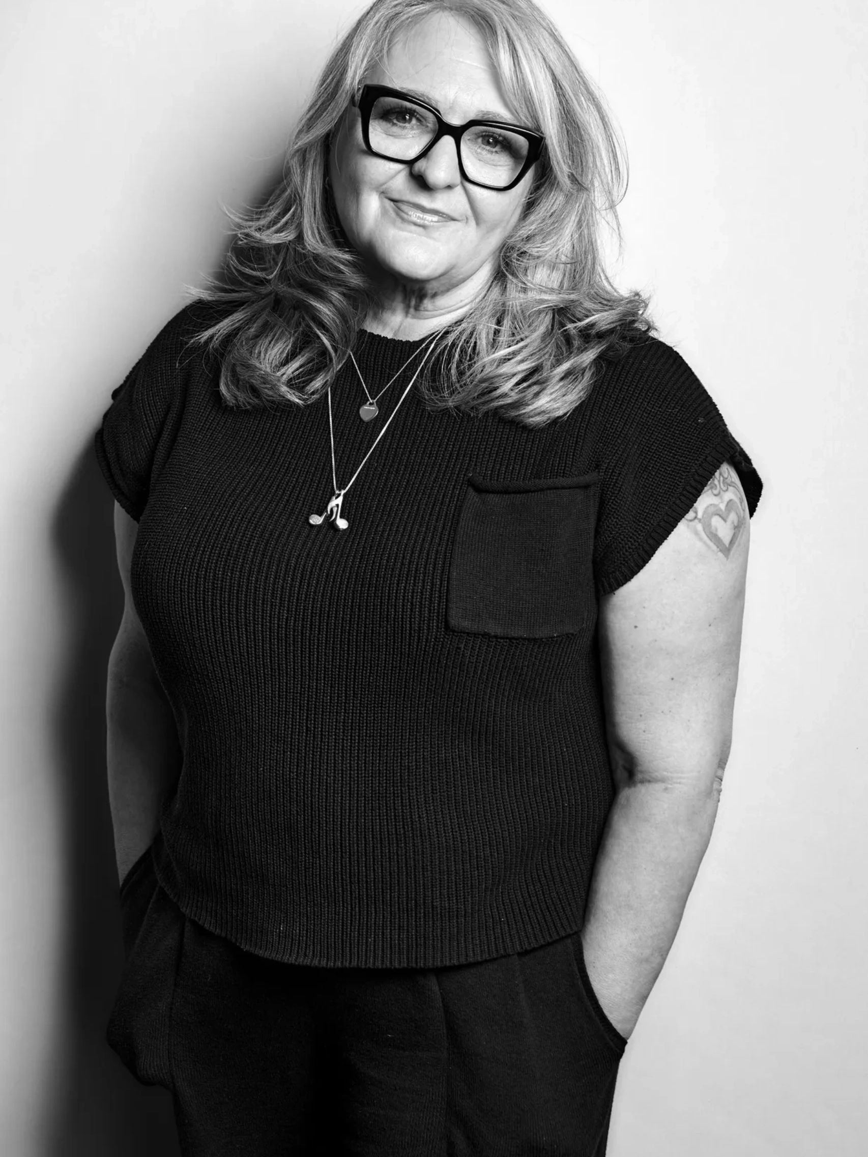 Smiling woman with glasses and tattoos, hands in pockets.