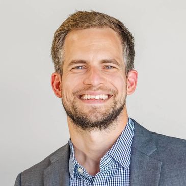 Smiling man in a blazer and checkered shirt against a plain background.