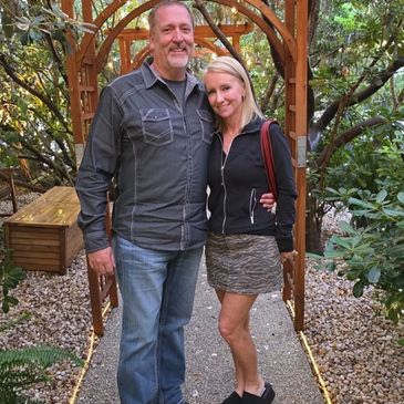 Couple smiling and posing under a wooden garden arch surrounded by greenery.
