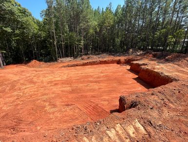 A large, freshly excavated plot of red soil surrounded by trees under a clear blue sky.