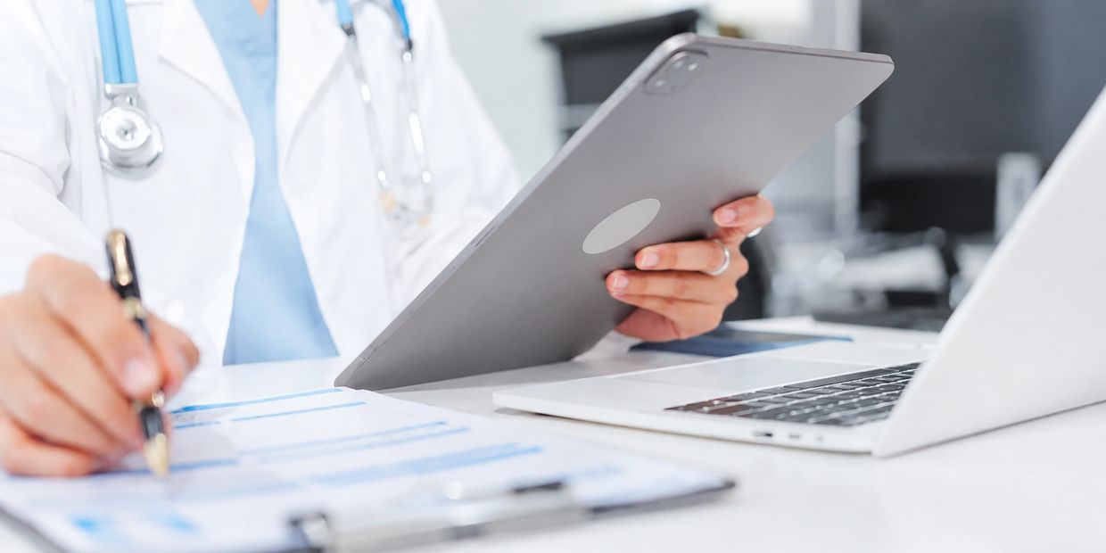 Doctor using tablet and laptop while writing on paperwork.