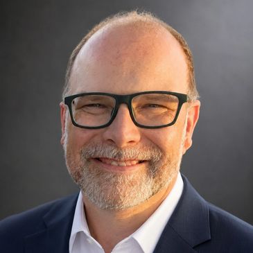 Smiling man with glasses in suit against a dark background.