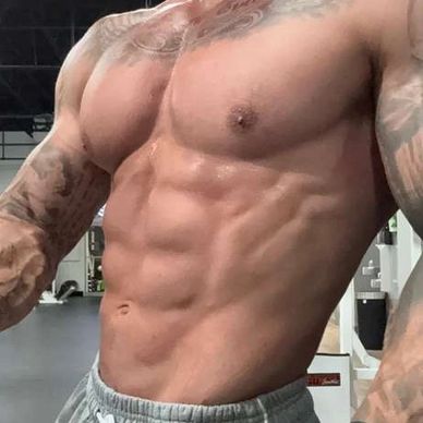 Muscular man with tattoos posing shirtless in a gym, wearing wrist wraps and gray sweatpants.