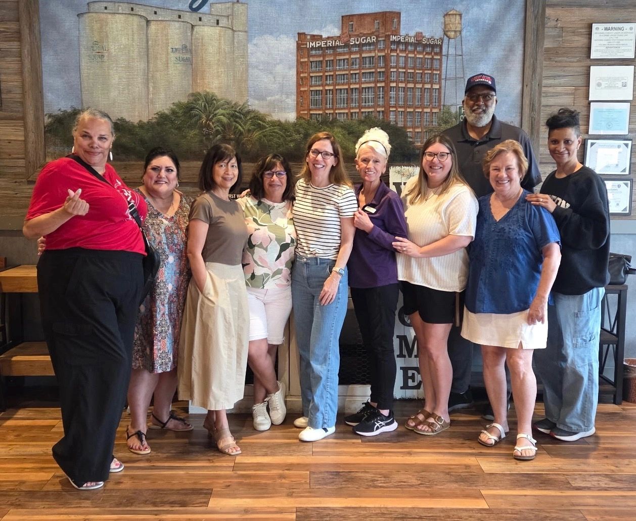 A diverse group of people smiling indoors with a mural of industrial buildings behind.
