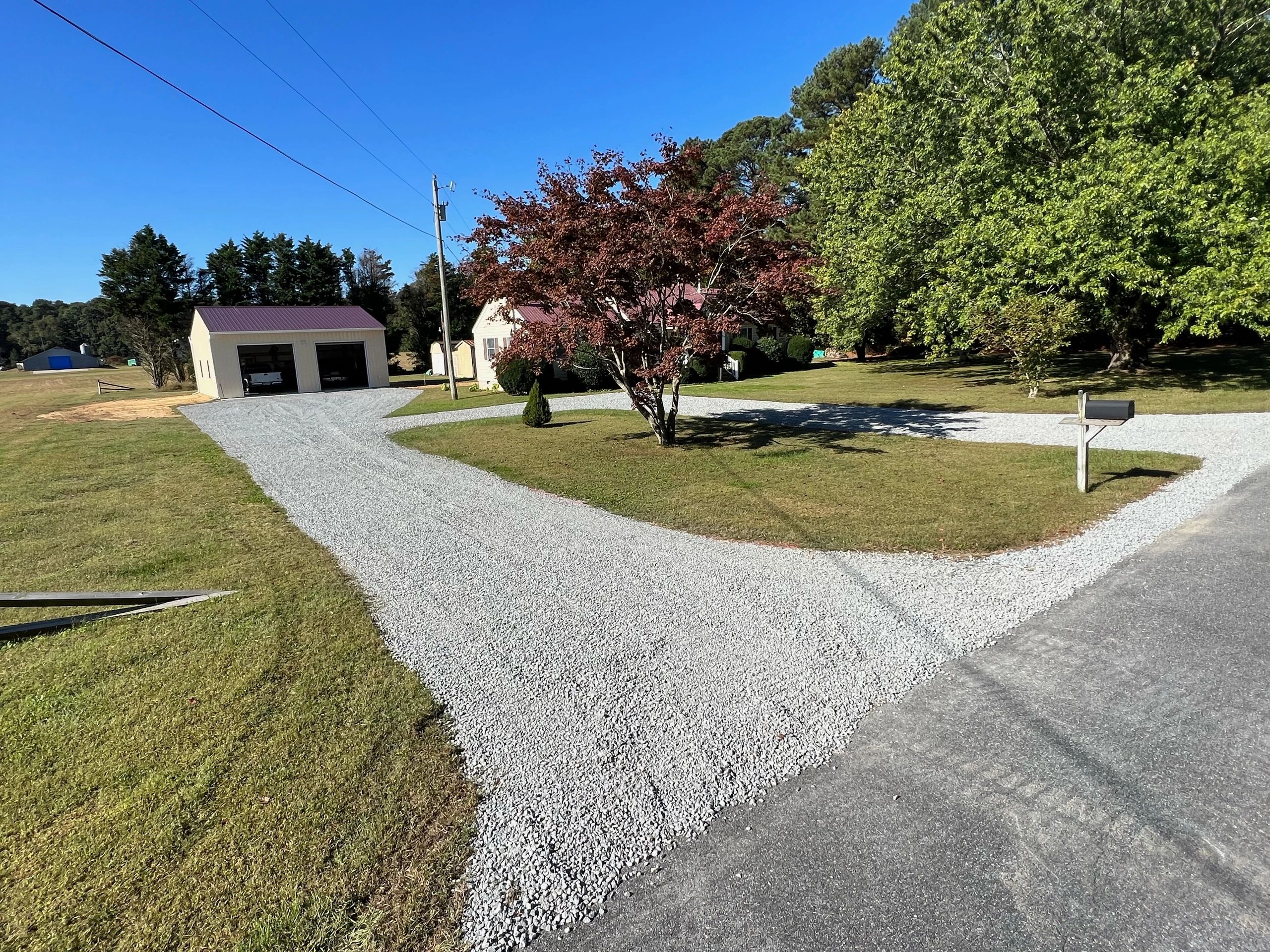 A rural house with a gravel driveway and lush green trees under a clear blue sky.